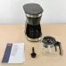 Lifestyle view of DeLonghi ICM12011 Active Line Coffee Maker with carafe, scoop, and manual on wooden surface.