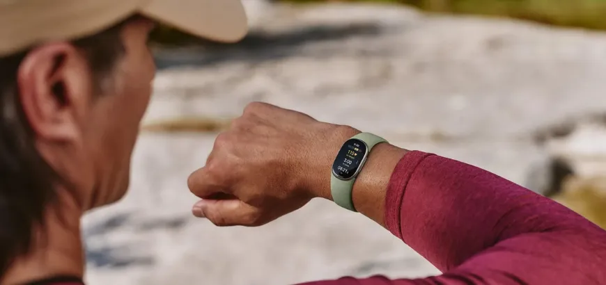 Man in a beige cap checking a green-banded Google Fitbit Edge on his wrist during an outdoor workout, with heart rate and timer on screen
