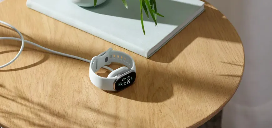 Olive-banded Google Fitbit Edge charging on a round wooden table, next to a book and a green plant