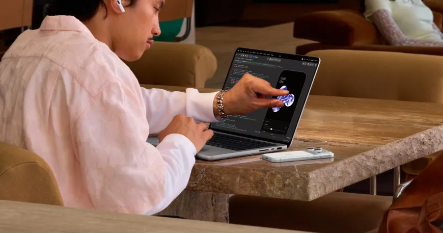Side view of a person wearing AirPods seated at a stone coffee table, interacting with a touchscreen laptop while working.